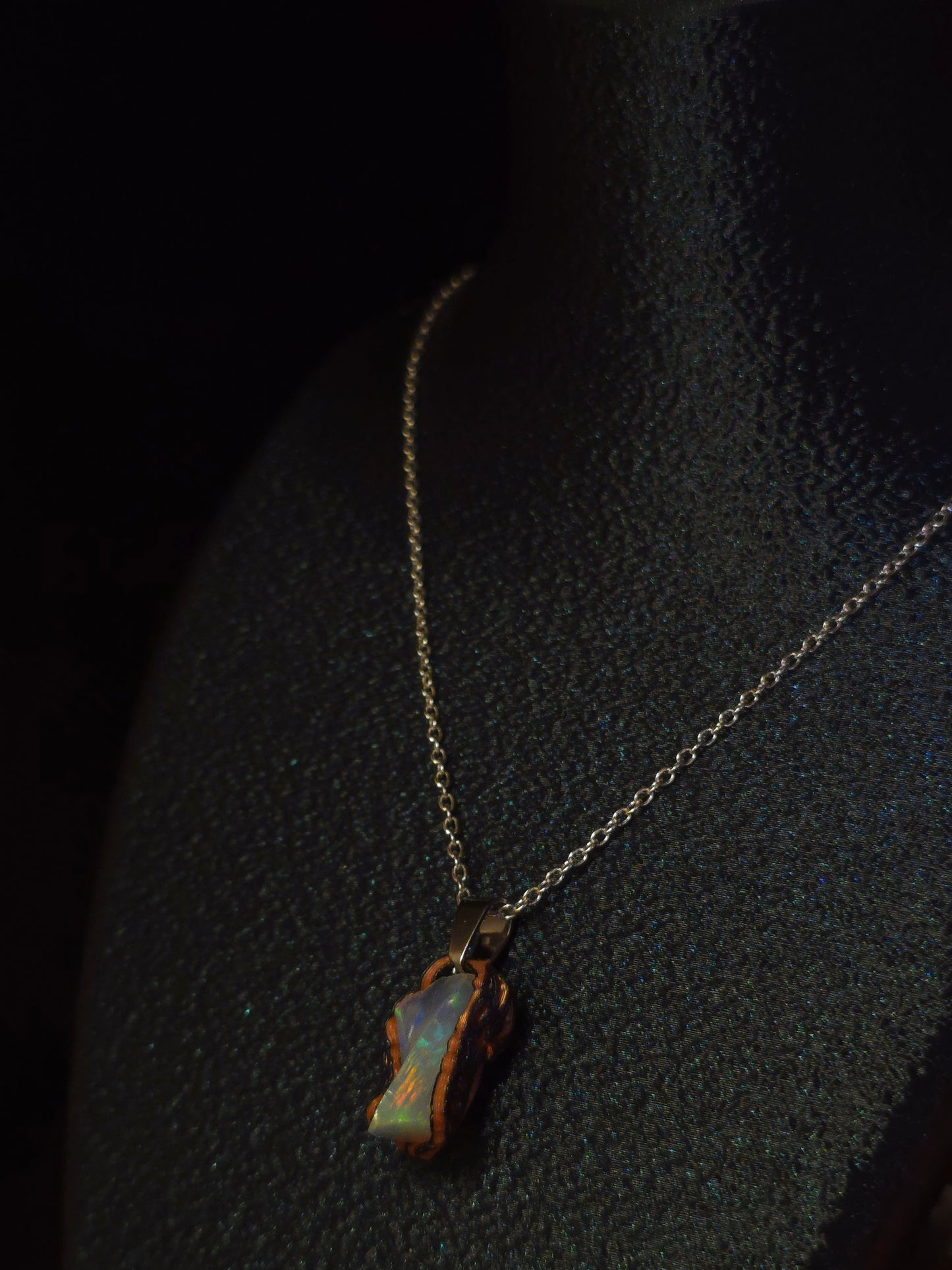A delicate gold chain necklace with an irregular, organic-shaped pendant made of iridescent opal showing blue, green, and orange hues, displayed on a textured black surface.