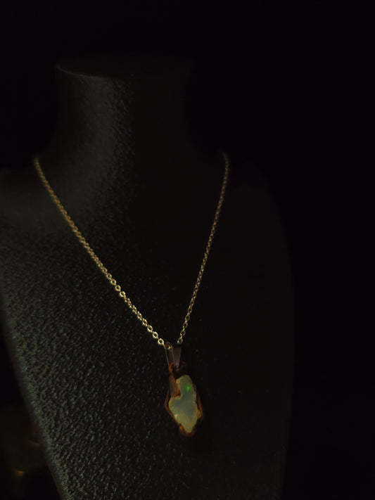 A delicate gold chain necklace with a small, irregularly shaped iridescent pendant resting on a textured black display stand against a dark background.