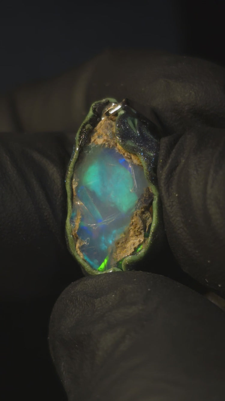 Video: raw uncut Ethiopian Welo opal pendant. Colour: green and blue, strong. Body: clear, light blue. Held in a gloved hand against a dark background.