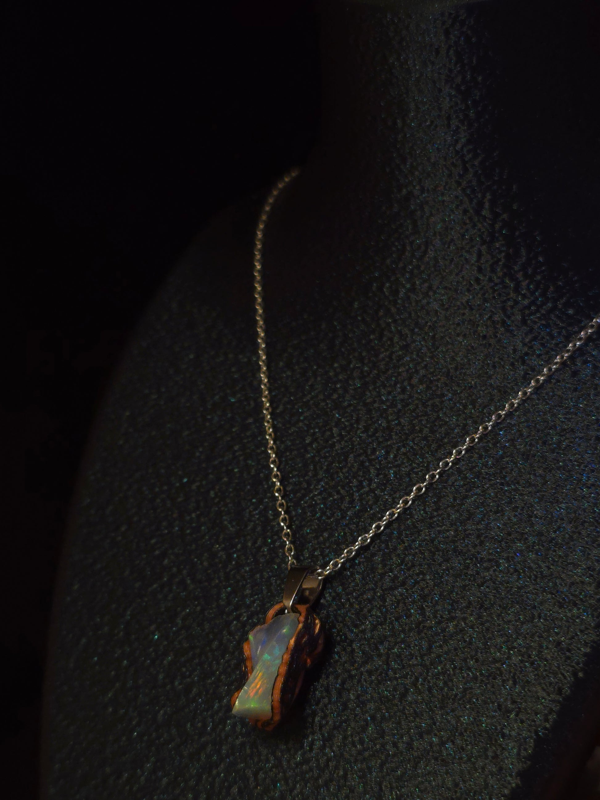A delicate gold chain necklace with an irregular, organic-shaped pendant made of iridescent opal showing blue, green, and orange hues, displayed on a textured black surface.