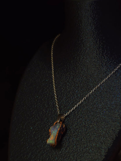 A delicate gold chain necklace with an irregular, organic-shaped pendant made of iridescent opal showing blue, green, and orange hues, displayed on a textured black surface.