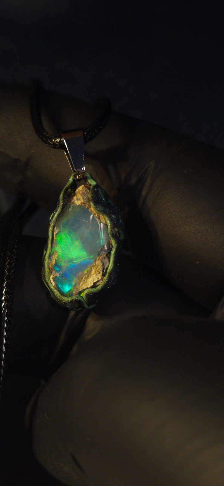 Raw uncut Ethiopian Welo opal pendant. Colour: green and blue, strong. Body: clear, light blue. Held in a gloved hand against a dark background.