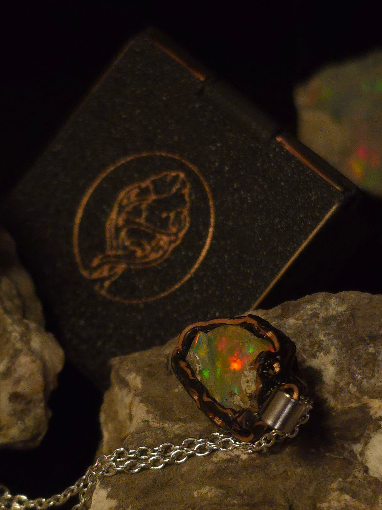 A raw opal pendant necklace with a silver chain and a dark metal bezel, displayed on rocks next to its black presentation box with a gold frog logo.