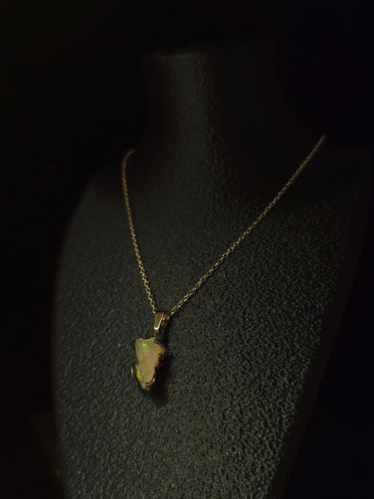 A gold necklace with a raw, irregular-shaped opal pendant displayed on a black textured jewelry bust against a dark background.