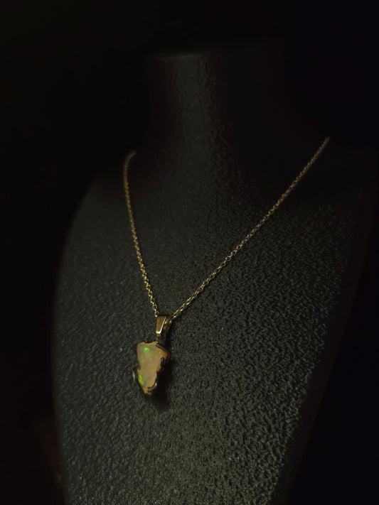 A gold necklace with a raw, irregular-shaped opal pendant displayed on a black textured jewelry bust against a dark background.