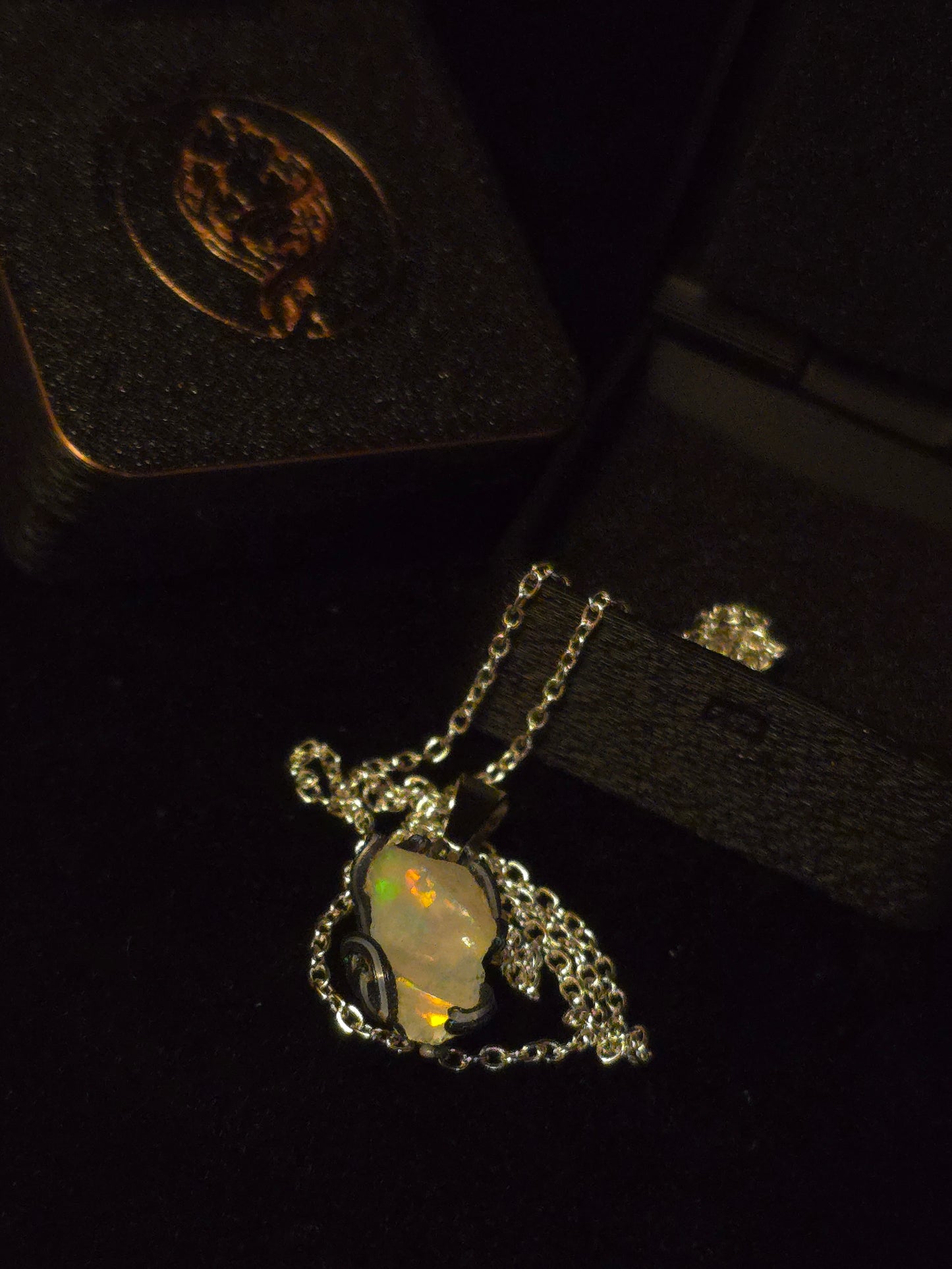 A silver-toned necklace with an opal pendant, displayed on a black velvet jewelry box next to its box lid.