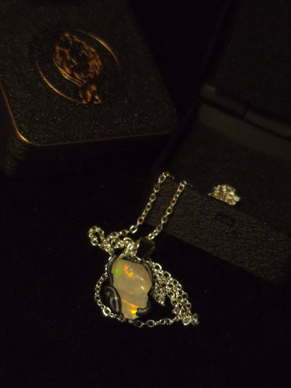A silver-toned necklace with an opal pendant, displayed on a black velvet jewelry box next to its box lid.