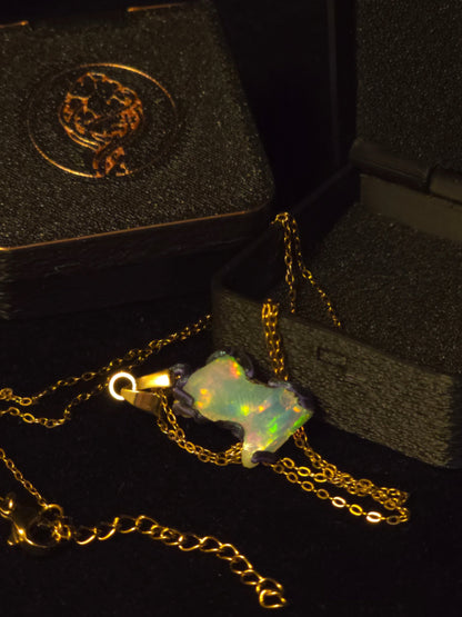 A gold chain necklace with a glowing opal pendant, displayed on a black surface next to its textured black presentation box.