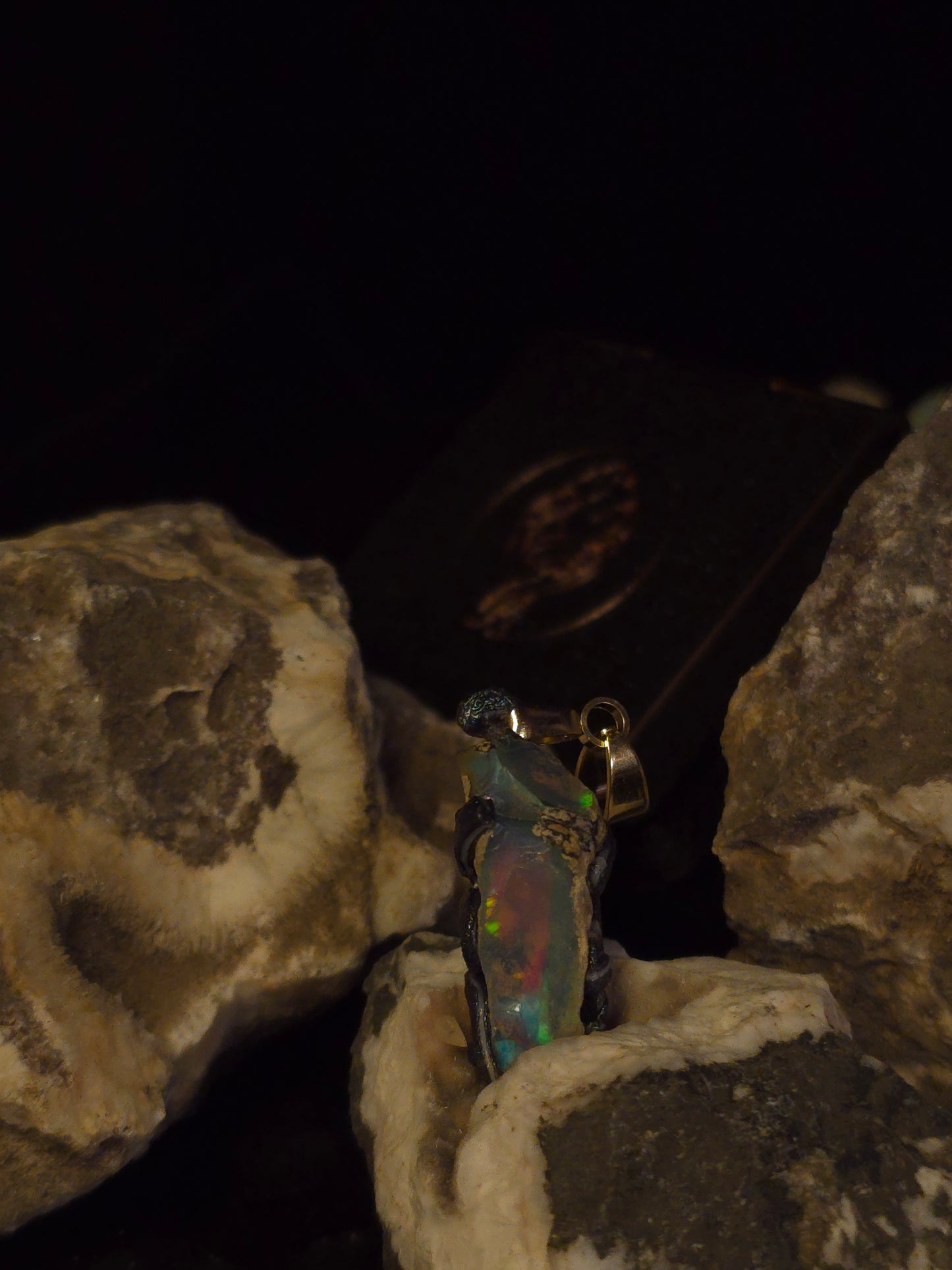 A small iridescent pendant or charm with a gold-toned loop, resting on rough natural rocks against a very dark background.