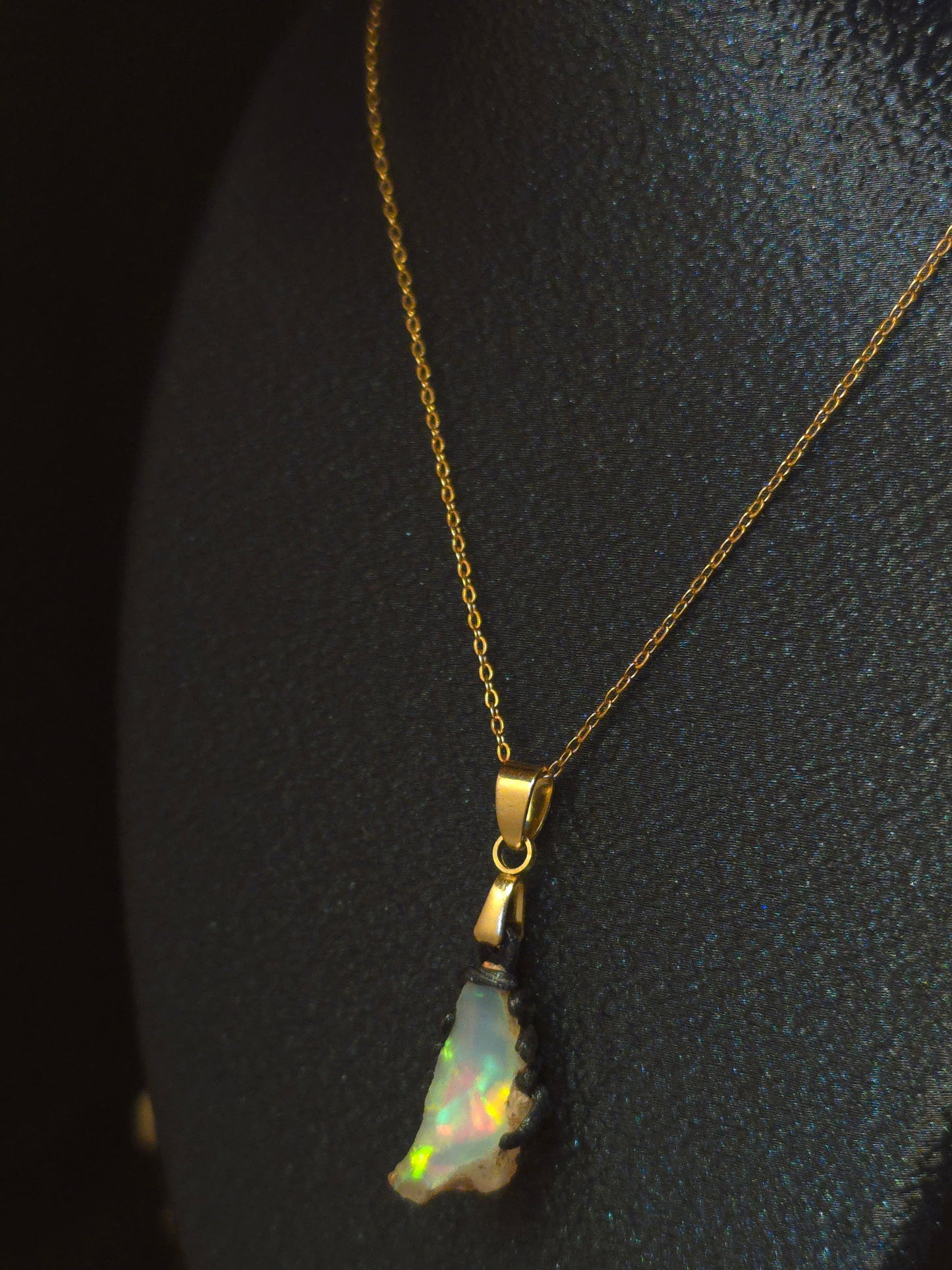 A gold chain necklace with a small iridescent opal pendant, displayed on a textured black surface.