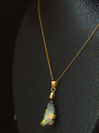 A gold chain necklace with a small iridescent opal pendant, displayed on a textured black surface.