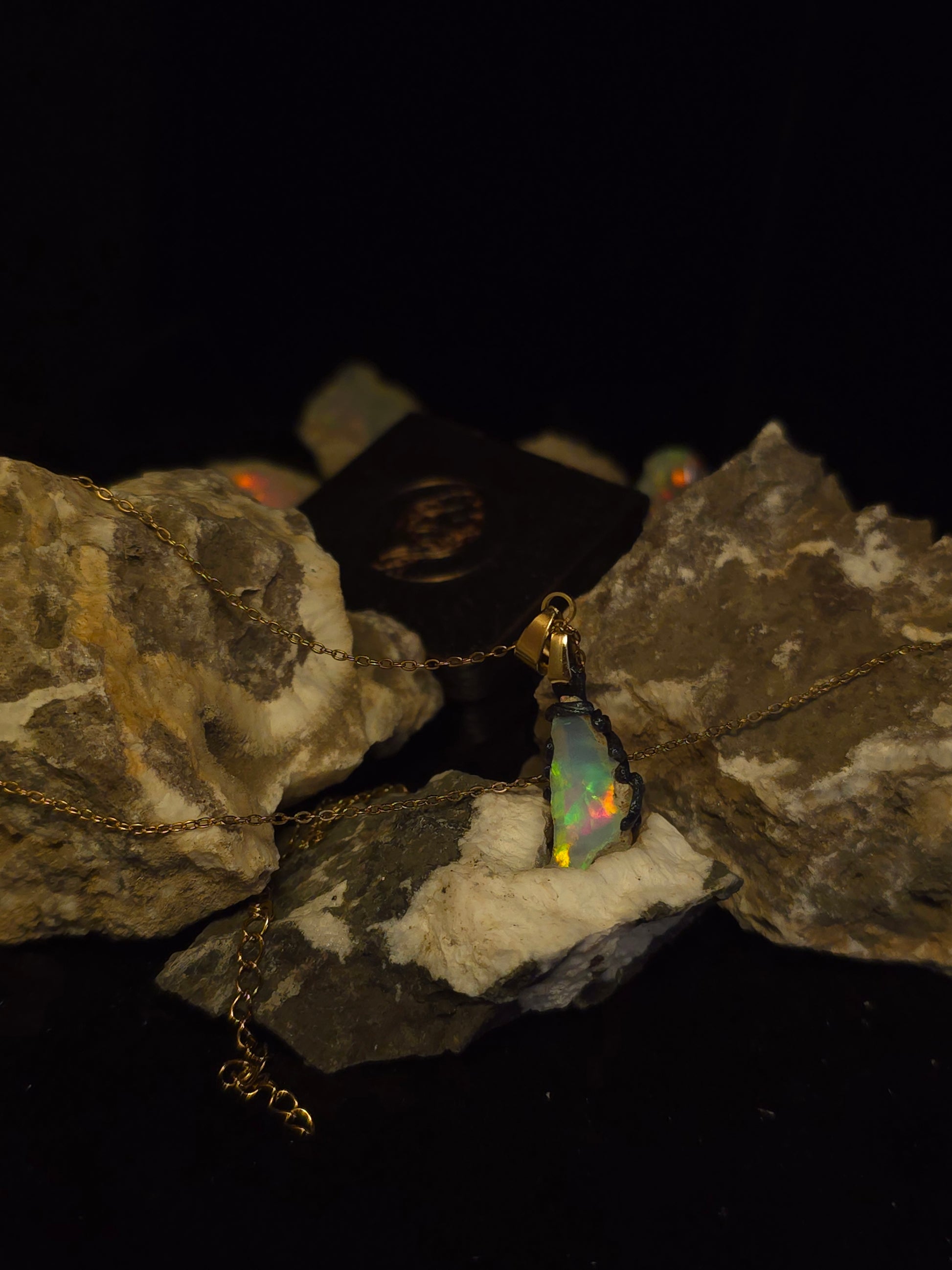 A gold chain necklace with a small iridescent opal pendant displayed on stacked natural rocks against a black background.
