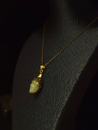 A gold pendant necklace with a small, irregularly shaped iridescent opal, displayed on a black jewelry bust against a dark background.