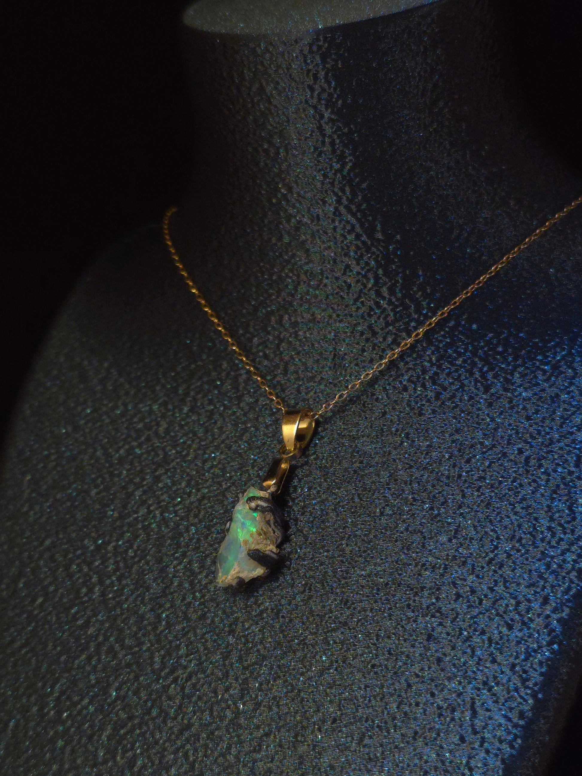 A gold chain necklace with a raw, irregular opal pendant displayed on a textured black mannequin bust.