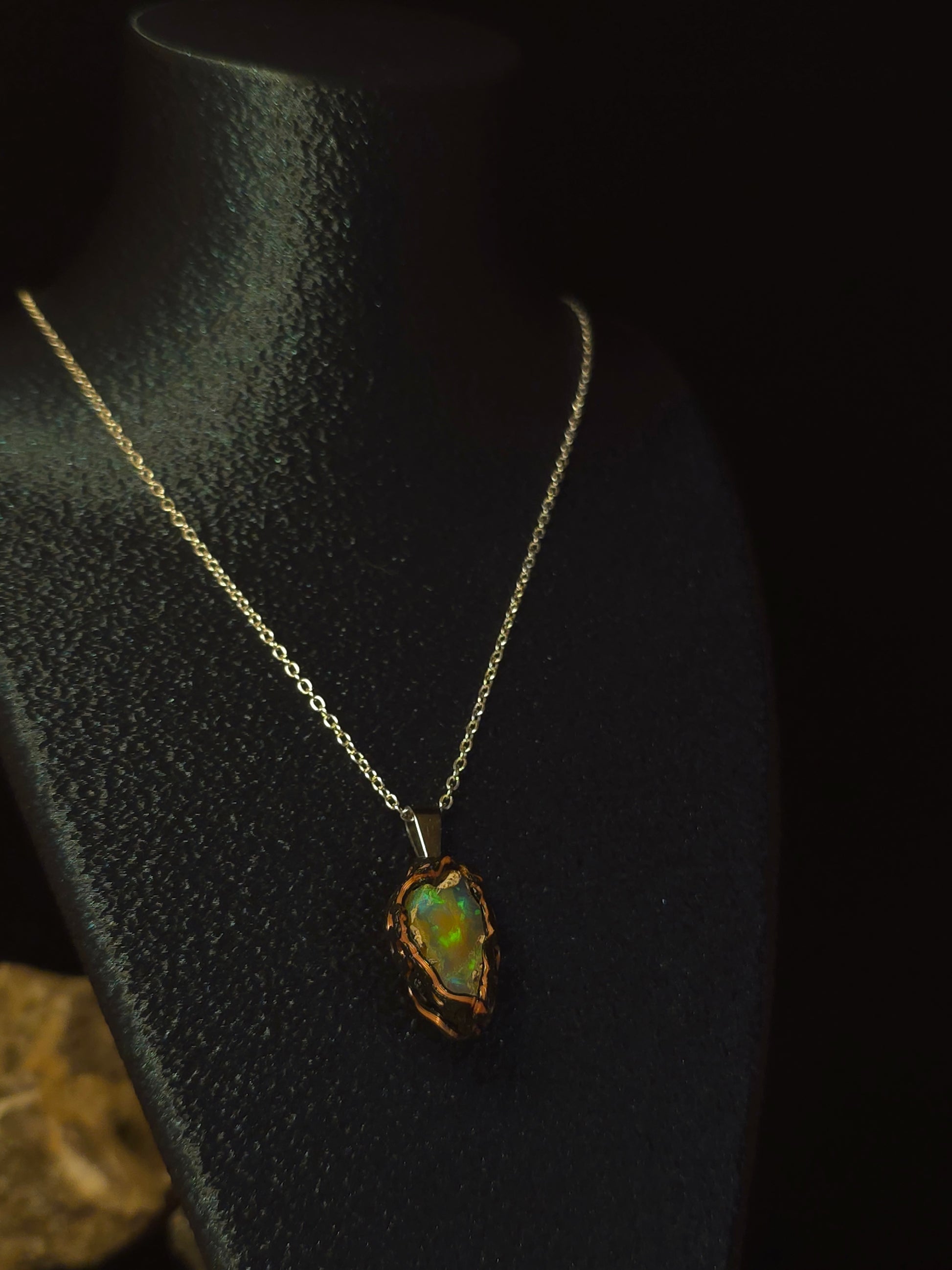 A gold chain necklace with a raw, irregular opal pendant displayed on a black jewelry bust against a dark background.