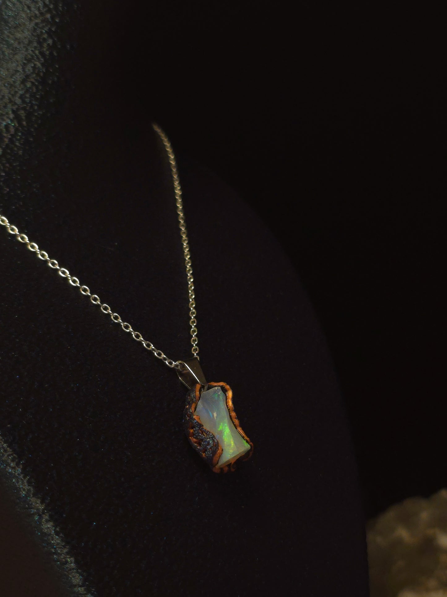 A delicate stainless steel chain necklace featuring a small, irregularly shaped iridescent opal pendant with a rustic, rough stone border, displayed on a dark velvet surface.