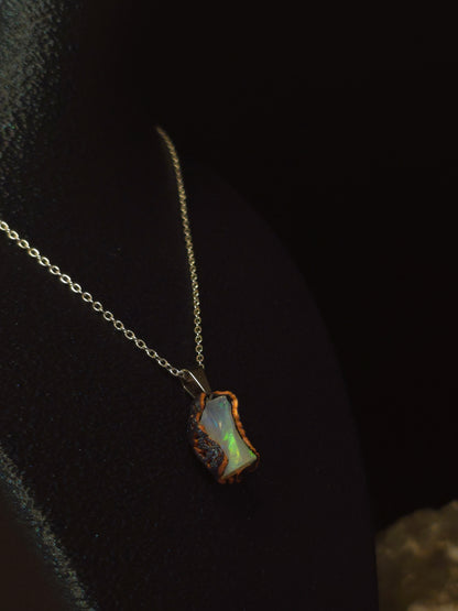 A delicate stainless steel chain necklace featuring a small, irregularly shaped iridescent opal pendant with a rustic, rough stone border, displayed on a dark velvet surface.