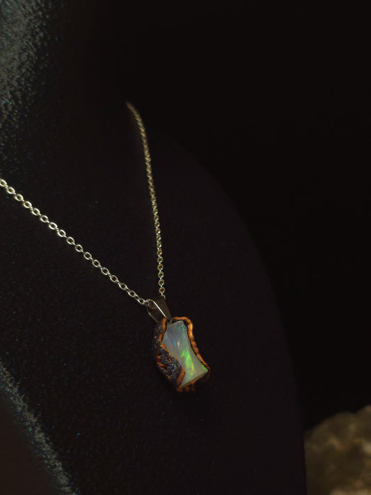 A delicate stainless steel chain necklace featuring a small, irregularly shaped iridescent opal pendant with a rustic, rough stone border, displayed on a dark velvet surface.