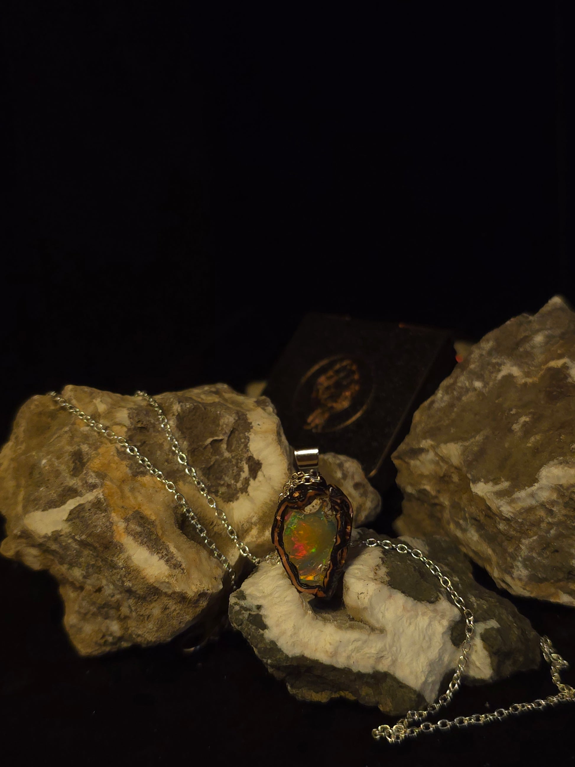 A silver chain necklace with a teardrop-shaped opal pendant, displayed on textured rocks against a black background.
