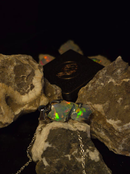 A silver-toned necklace with a pendant featuring a highly iridescent, multicolored opal showing green, orange, and blue flashes, displayed on textured rock formations against a black background.