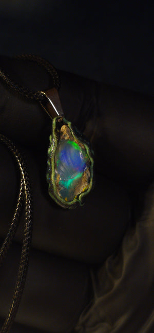 Raw uncut Ethiopian Welo opal pendant. Colour: green and blue, strong. Body: clear, light blue. Held in a gloved hand against a dark background.