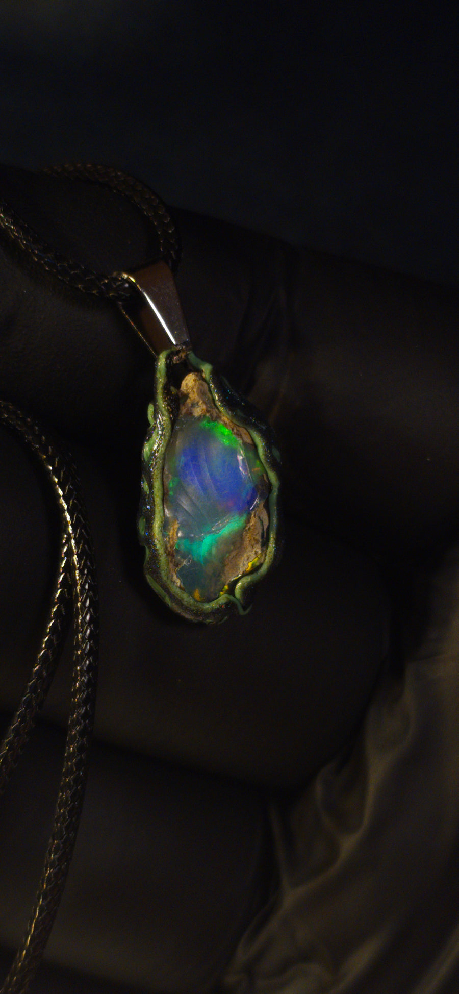 Raw uncut Ethiopian Welo opal pendant. Colour: green and blue, strong. Body: clear, light blue. Held in a gloved hand against a dark background.