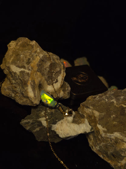 A necklace with a small glowing neon yellow-green opal pendant set in a gold-toned frame, displayed on rough rocks against a black background.