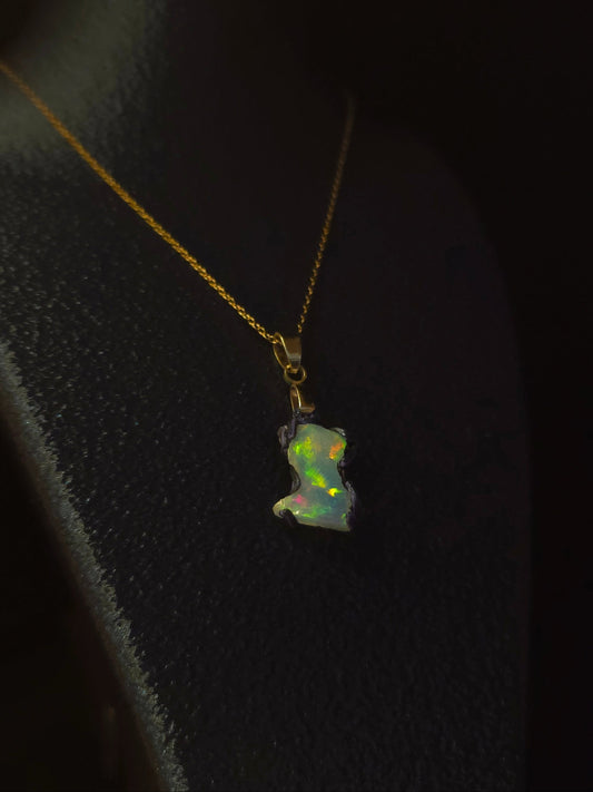 A delicate gold necklace with a small, irregularly shaped opal pendant, displayed on a black textured jewelry bust against a dark background.