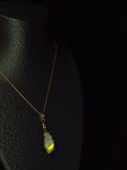 A delicate gold chain necklace with a small, irregularly shaped iridescent  opal pendant, displayed on a black textured surface against a dark background.