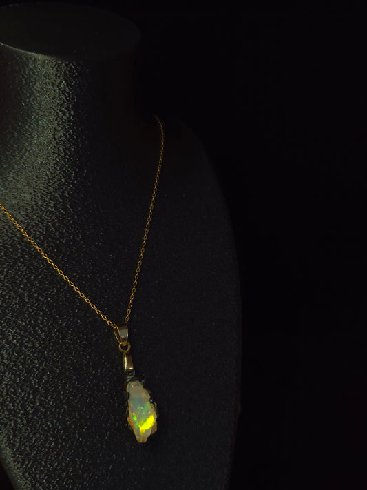A delicate gold chain necklace with a small, irregularly shaped iridescent  opal pendant, displayed on a black textured surface against a dark background.