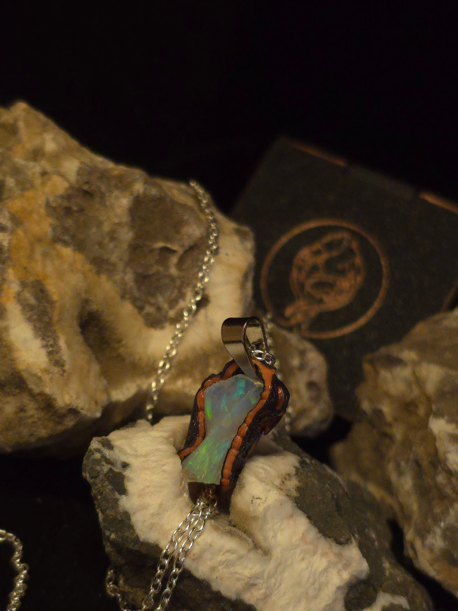 A silver chain necklace with a pendant made from an iridescent green-blue opal with a dark brown textured border, displayed on textured rocks against a black background.