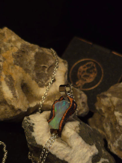 A silver chain necklace with a pendant made from an iridescent green-blue opal with a dark brown textured border, displayed on textured rocks against a black background.