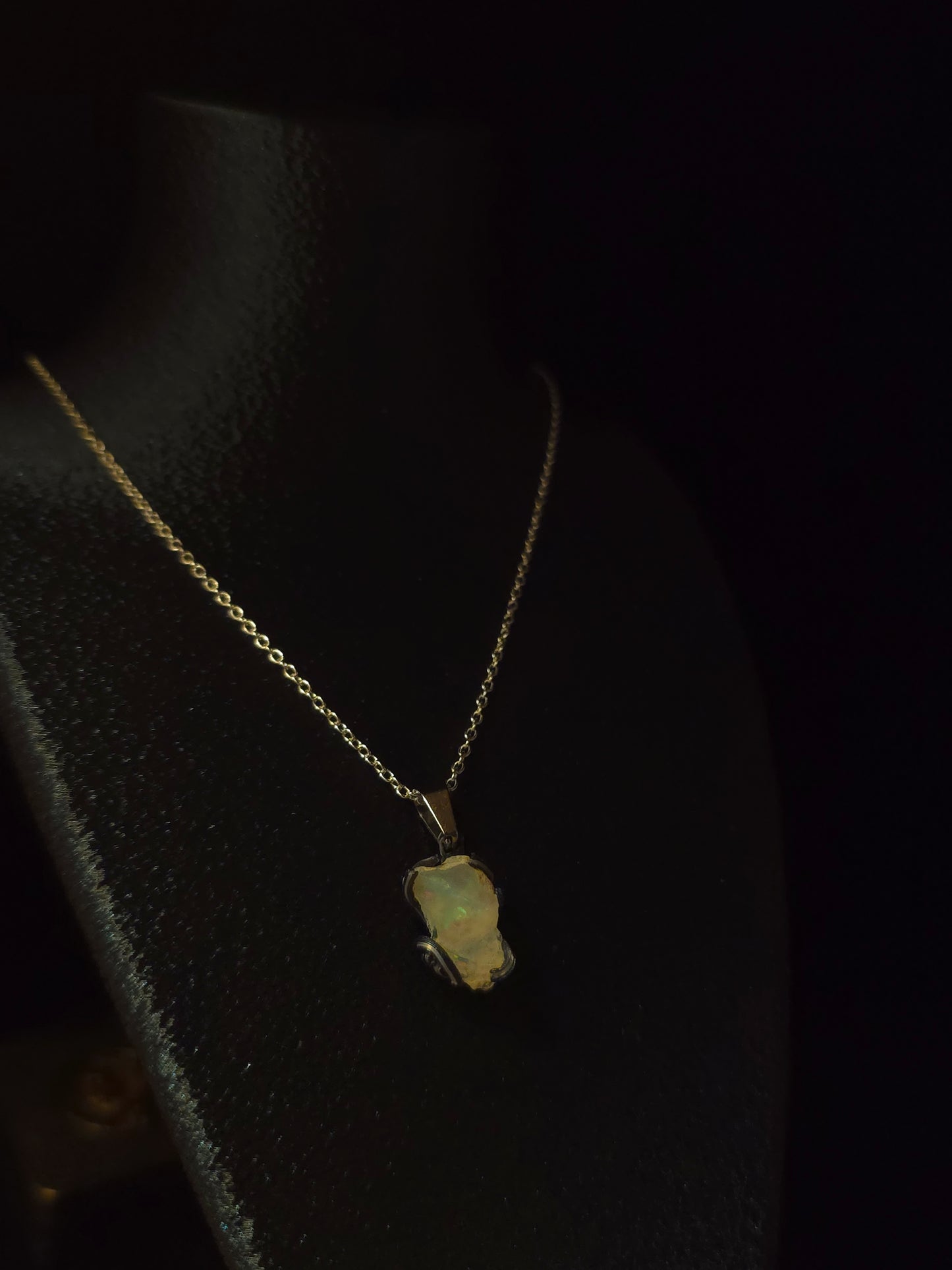 A delicate gold chain necklace with a small, irregular pale green opal pendant, displayed on a black jewelry bust against a dark background.