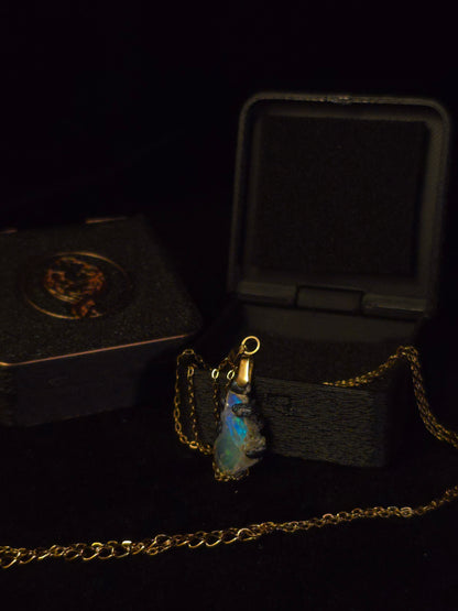 A gold chain necklace with a raw, irregular opal pendant, displayed next to its black presentation box on a black background.