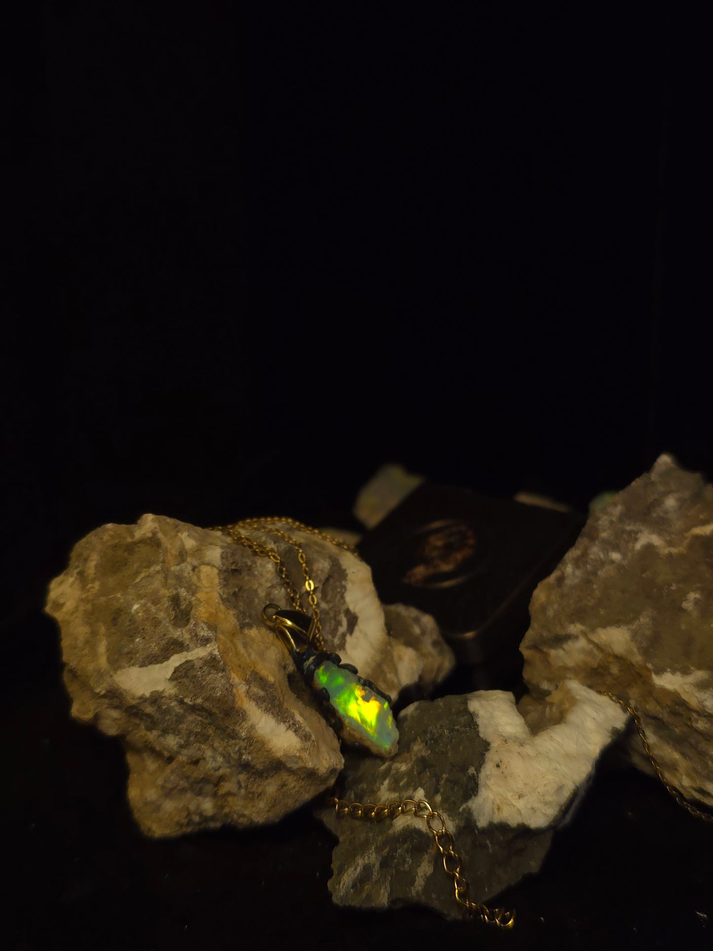 A gold chain necklace with a small iridescent opal pendant resting on rough rocks against a solid black background.