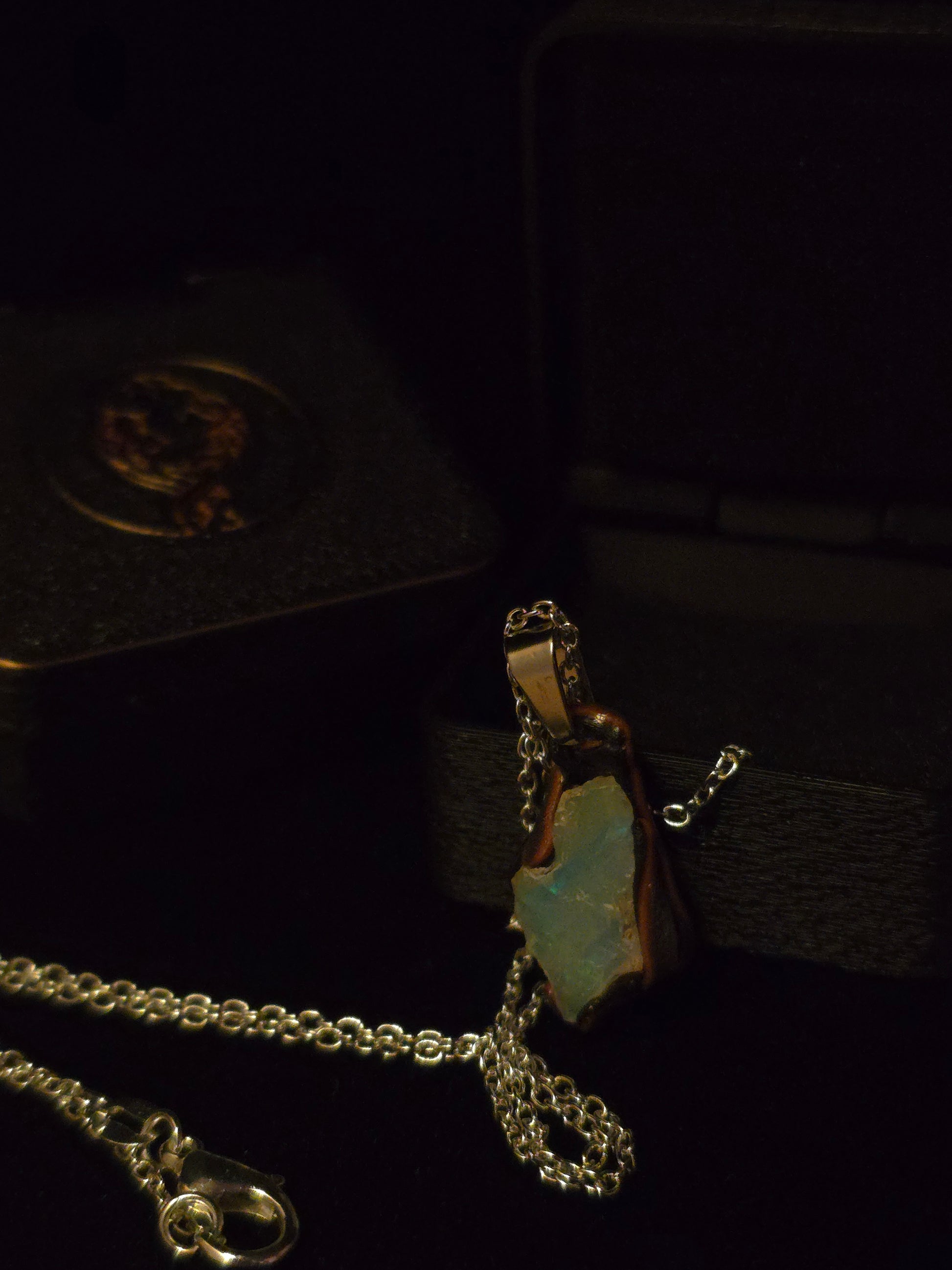 A silver-toned chain necklace with a pendant featuring an irregular light blue-green opal set in a gold-toned frame, displayed on a dark velvet jewelry box.