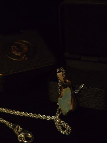 A silver-toned chain necklace with a pendant featuring an irregular light blue-green opal set in a gold-toned frame, displayed on a dark velvet jewelry box.