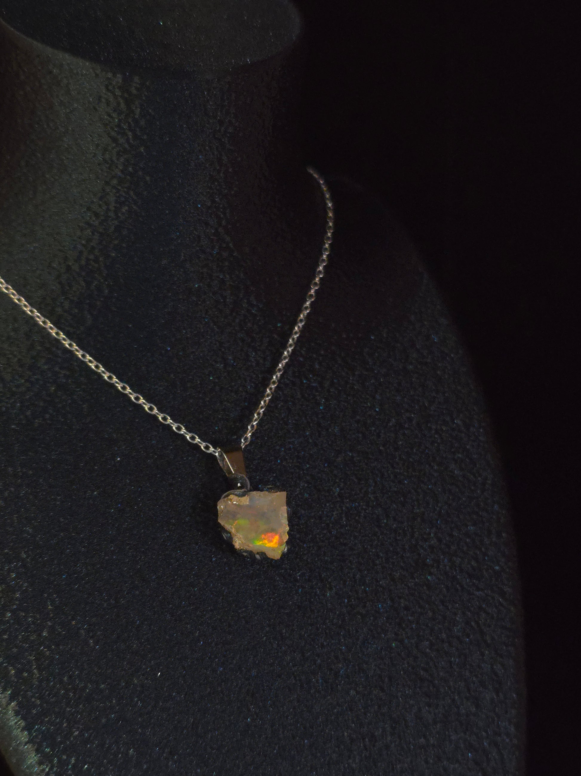 A silver chain necklace with a small, rough, irregularly shaped opal pendant, displayed on a black jewelry bust against a dark background.