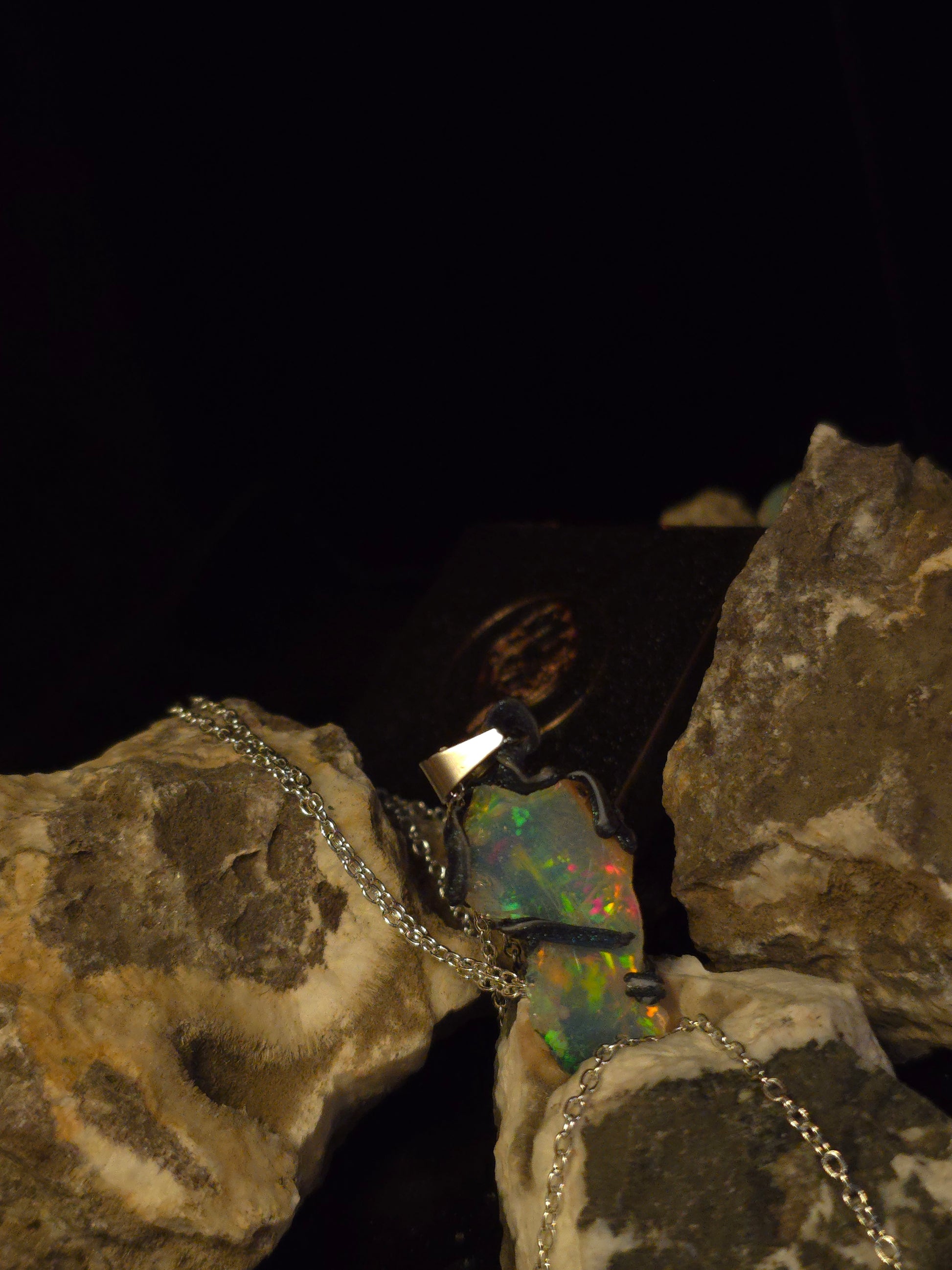 A silver chain necklace with a pendant featuring a colorful opal, displayed on rough rocks against a dark background.