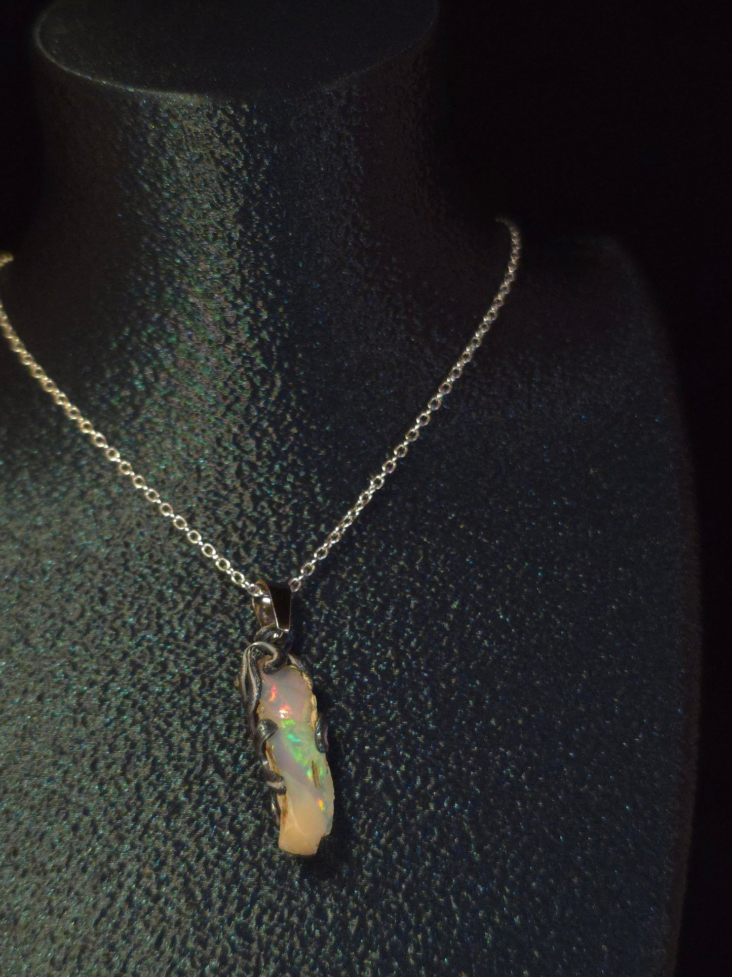 A delicate silver chain necklace with a small, irregular opal pendant, displayed on a black textured jewelry bust.