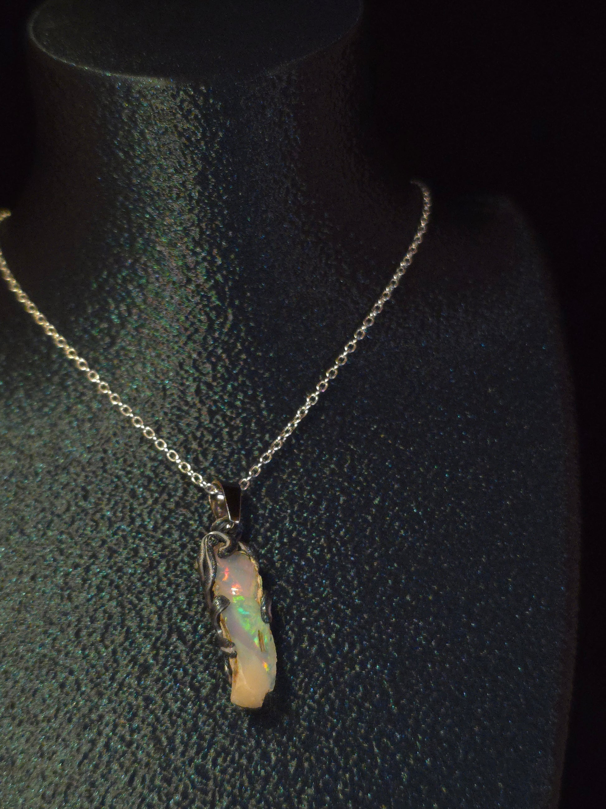 A delicate silver chain necklace with a small, irregular opal pendant, displayed on a black textured jewelry bust.