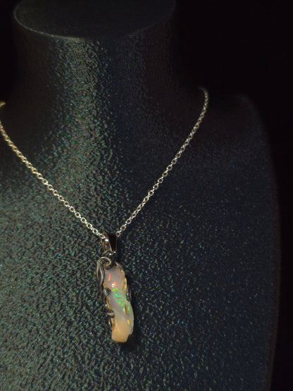 A delicate silver chain necklace with a small, irregular opal pendant, displayed on a black textured jewelry bust.