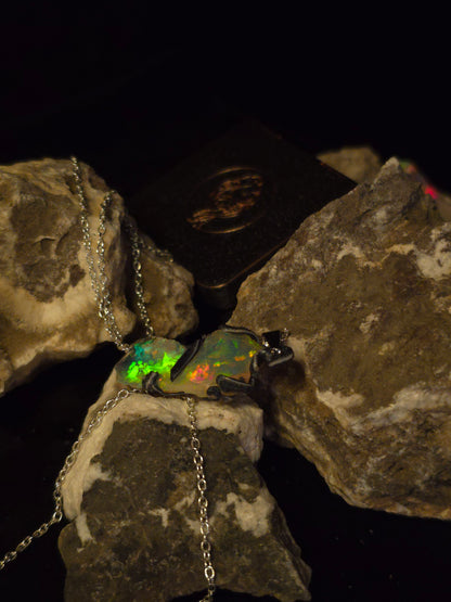 A silver chain necklace with an opal pendant displayed on rough rocks against a black background. The opal shows iridescent colors including green, orange, and blue.