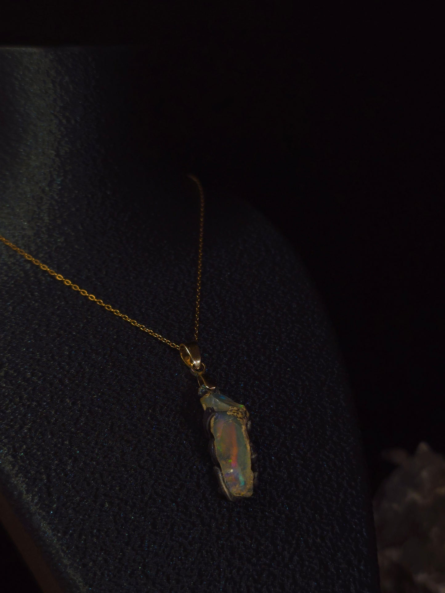 A gold chain necklace with a raw, irregular opal pendant displayed on a black jewelry bust against a dark background.