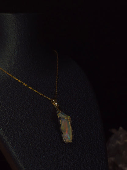 A gold chain necklace with a raw, irregular opal pendant displayed on a black jewelry bust against a dark background.