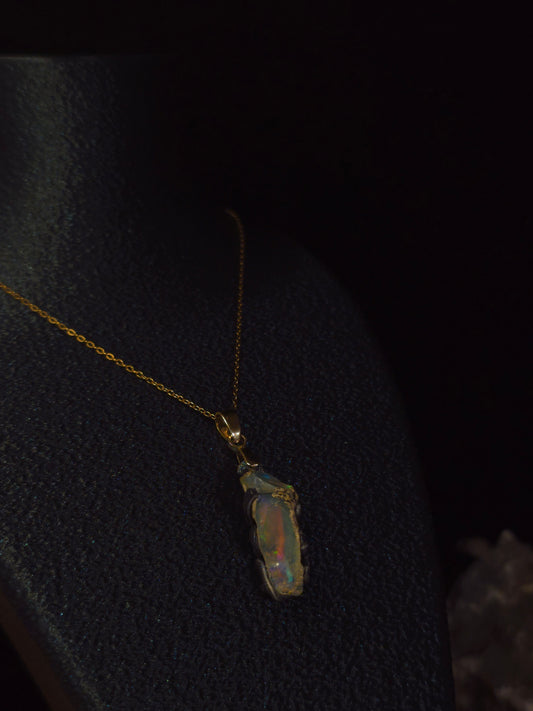 A gold chain necklace with a raw, irregular opal pendant displayed on a black jewelry bust against a dark background.