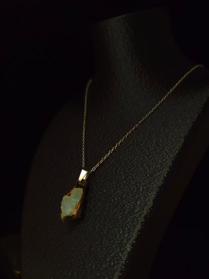 A gold chain necklace with a raw opal pendant displayed on a black textured mannequin bust against a dark background.
