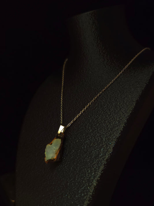 A gold chain necklace with a raw opal pendant displayed on a black textured mannequin bust against a dark background.