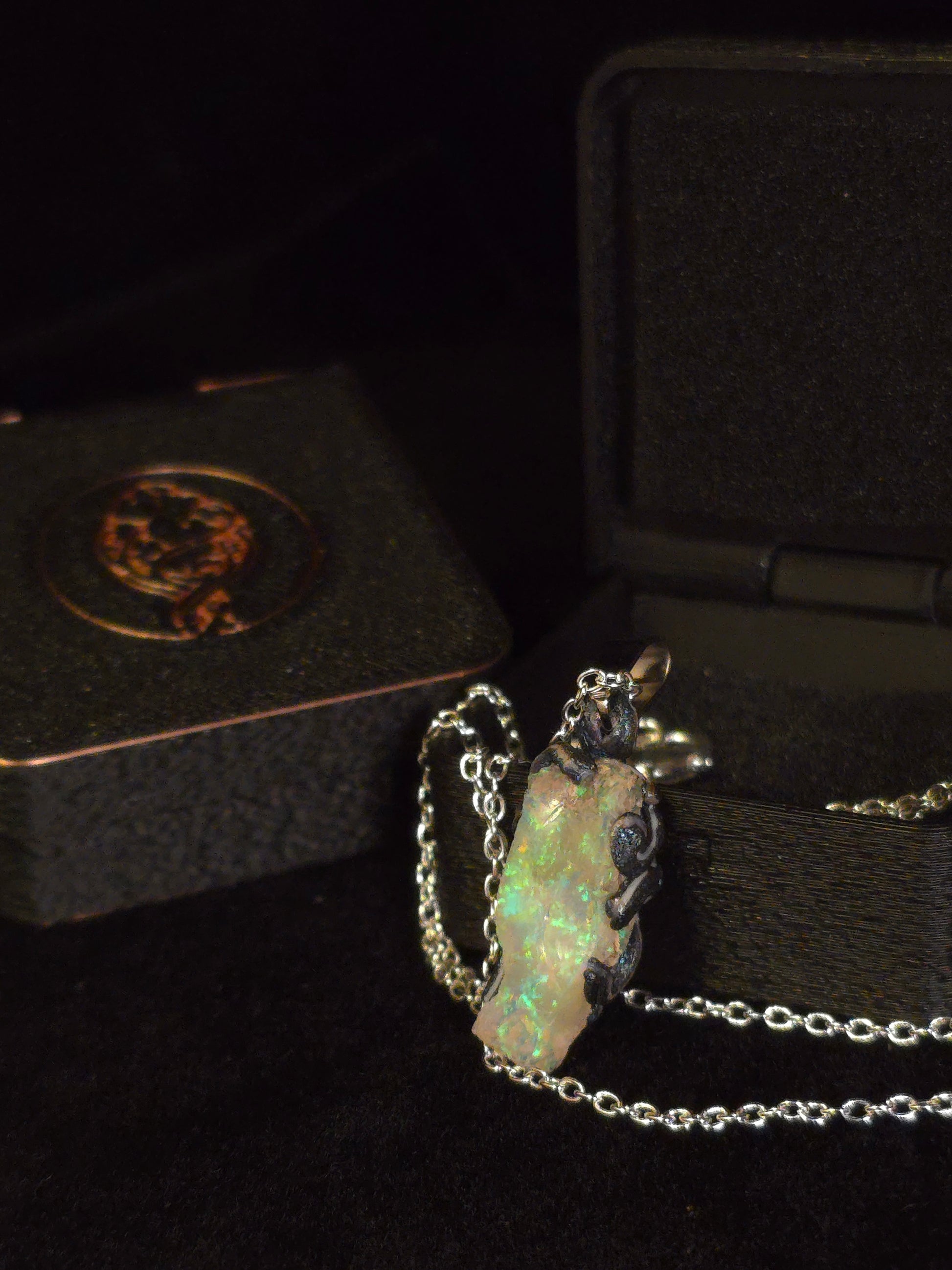 A silver chain necklace with a raw, irregular opal pendant displayed next to an open black jewelry box.
