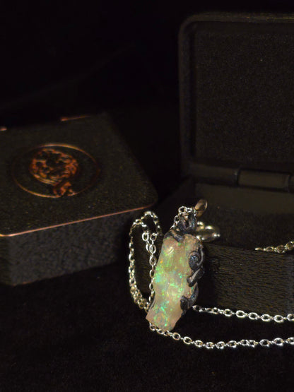A silver chain necklace with a raw, irregular opal pendant displayed next to an open black jewelry box.